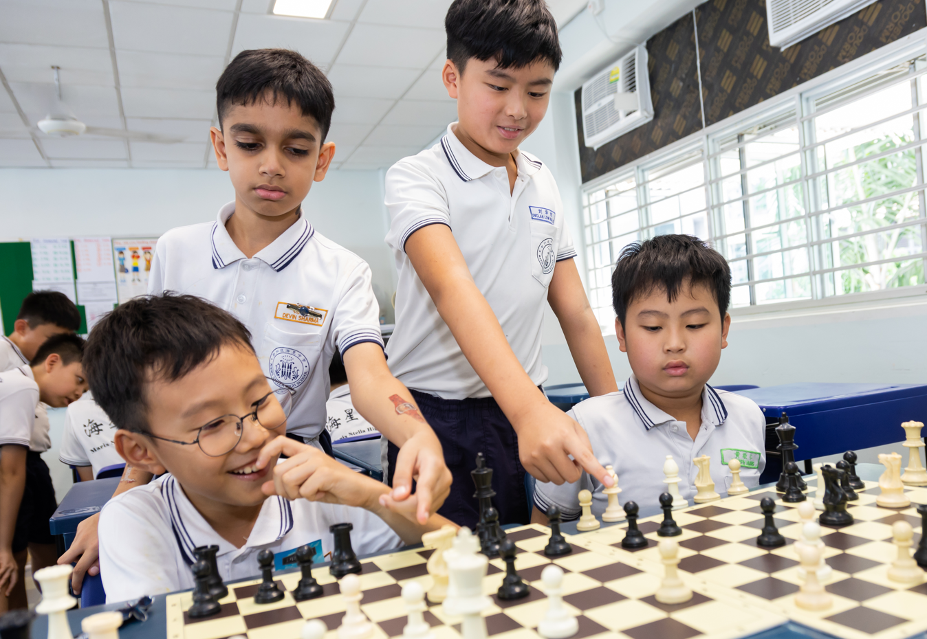 Two rows of children facing each other playing chess at tables with chess clocks in a classroom.
